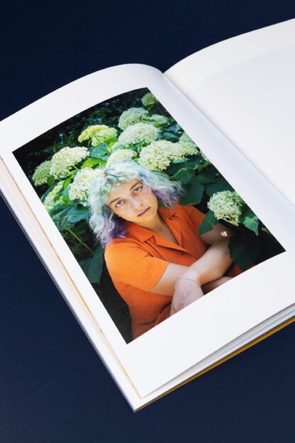 Strona książki ze zdjęciem młodej kobiety w pomarańczowym ubraniu wśród białych kwiatów.